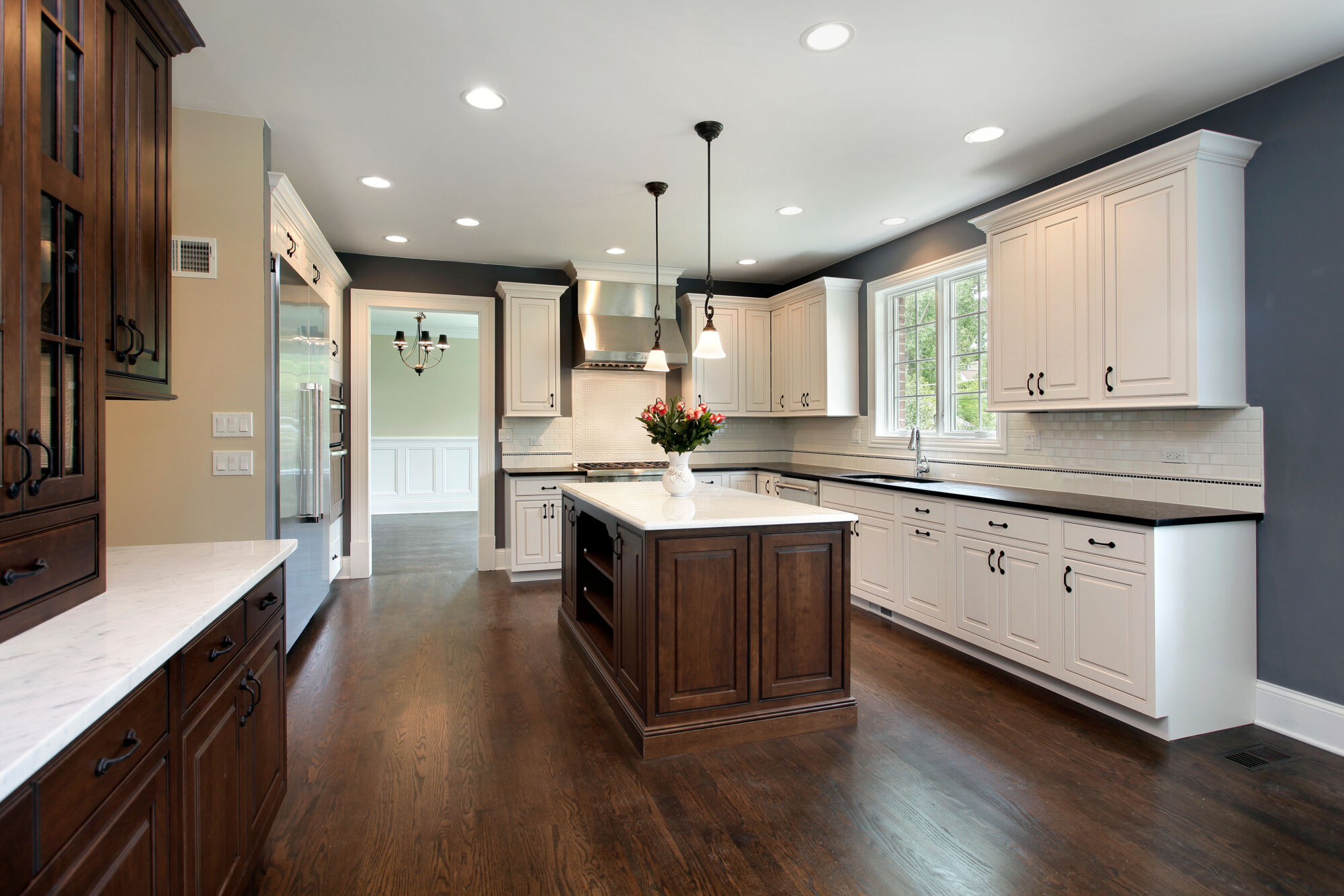 A spacious kitchen with raised panel white cabinets, a dark wood island with a marble countertop, and a dark wood floor. A vase of pink and red flowers sits on the island.
