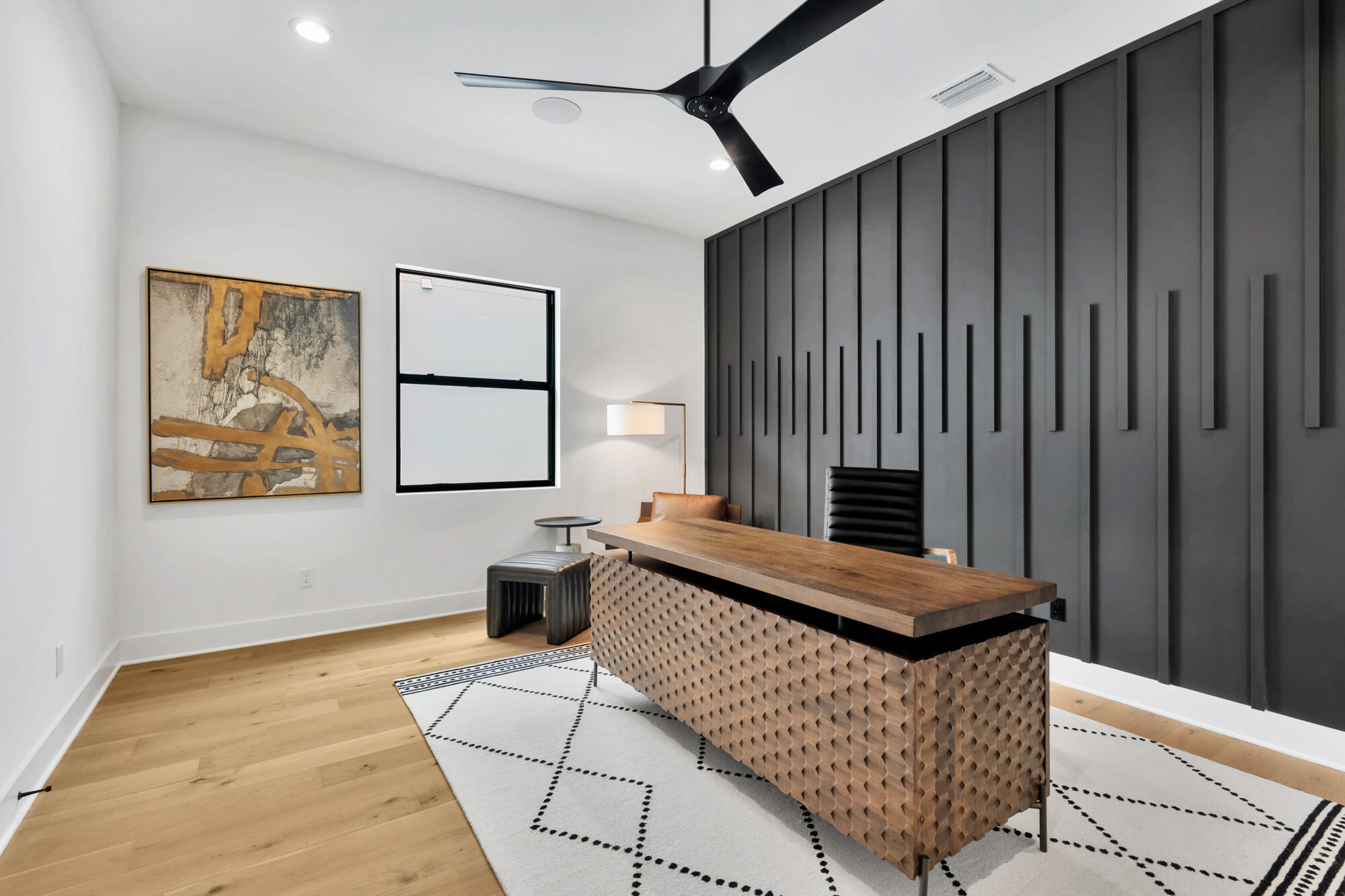 A modern office with a wooden desk, a patterned rug, abstract art on the wall, and a black slatted accent wall.